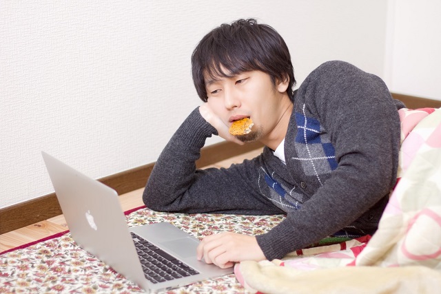 寝転がってせんべいを加えながらMacbookをいじっている男性